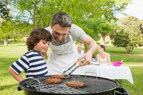 Man and son barbecuing with family in the background at park